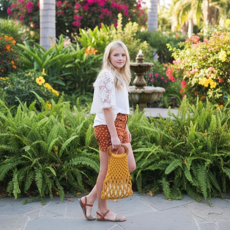 Peut inclure: Une personne marche dans un jardin, portant un sac en macram&eacute; jaune moutarde avec une poign&eacute;e en bois. Elle porte un haut en dentelle blanche, un short &agrave; pois marron et des sandales marron. L'arri&egrave;re-plan pr&eacute;sente une verdure luxuriante et une fontaine.