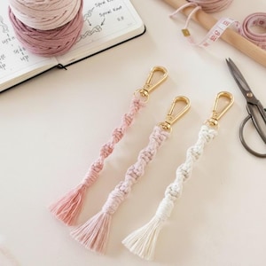 May include: Three macrame keychains in shades of pink and white, each with a gold-colored clasp and a tassel. The keychains are handmade with a spiral knot design. Crafting supplies, including yarn and scissors, are in the background.