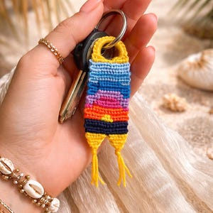 Op de afbeelding: Een handgemaakte sleutelhanger met een zonsondergangontwerp. De sleutelhanger is gemaakt van geweven draden in geel, blauw, roze, oranje en zwart. Het is bevestigd aan een zilveren sleutelhanger met autosleutels. Ongeveer 7 cm lang.