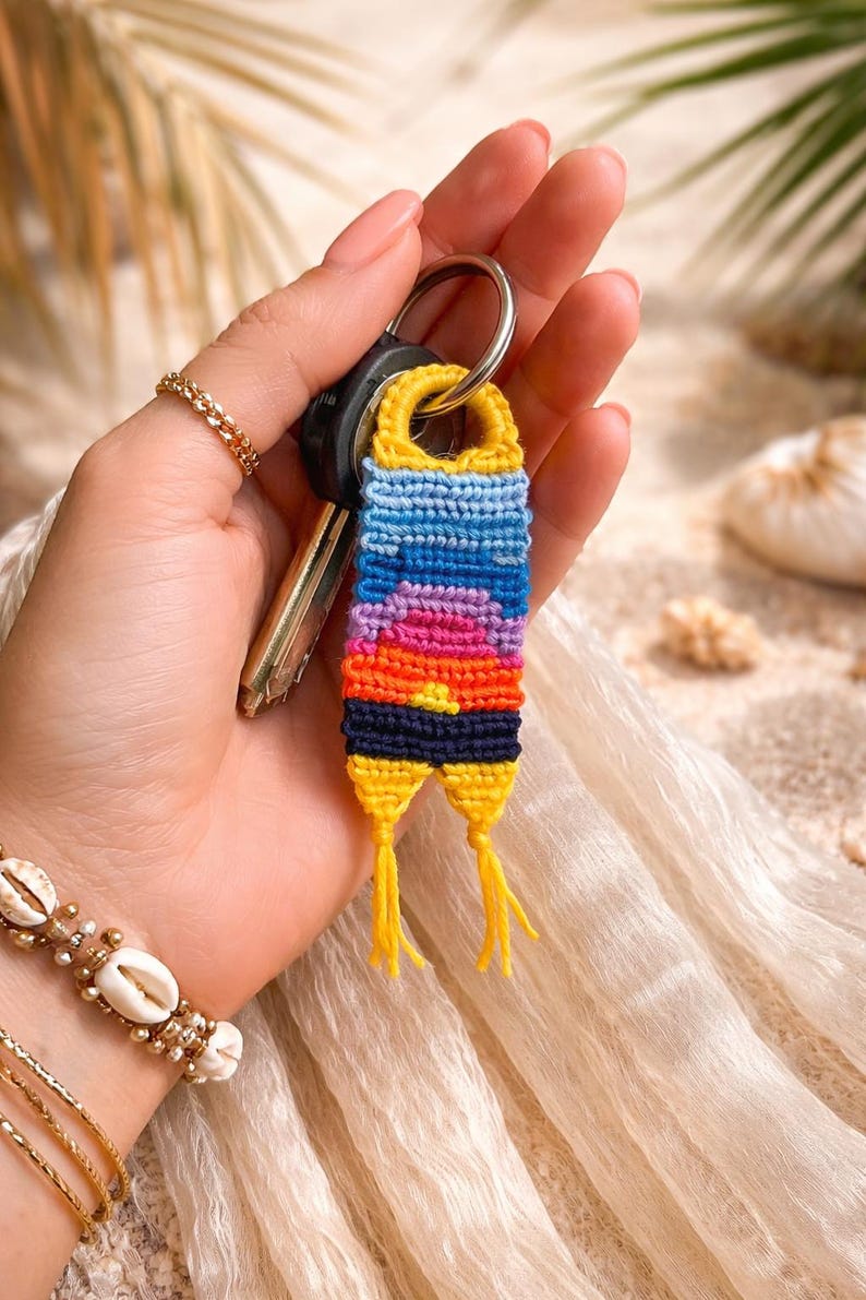 Op de afbeelding: Een handgemaakte sleutelhanger met een zonsondergangontwerp. De sleutelhanger is gemaakt van geweven draden in geel, blauw, roze, oranje en zwart. Het is bevestigd aan een zilveren sleutelhanger met autosleutels. Ongeveer 7 cm lang.