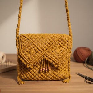 Peut inclure: Sac bandoulière en macramé jaune moutarde avec fermeture à rabat. Le sac présente un motif tissé avec des accents floraux et des détails de perles en bois. La bandoulière est également en macramé. Un accessoire élégant et unique.
