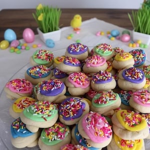 Può includere: Un piatto pieno di biscotti di zucchero glassati colorati. Ogni biscotto ha una glassa pastello diversa, tra cui rosa, viola, verde, giallo e blu, e sono ricoperti di zuccherini arcobaleno. Decorazioni a tema pasquale sullo sfondo.