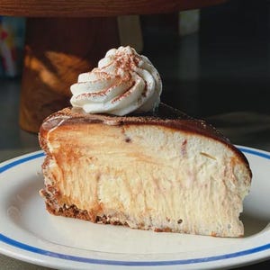 May include: A slice of cheesecake on a white plate with a blue rim. The cheesecake has a chocolate topping and is topped with whipped cream dusted with cocoa powder. The dessert is a sweet treat.