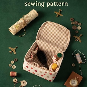 Op de afbeelding: Een naaipatroonkit voor jonge reizigers, met een reiskoffer met stippen en een gingham interieur. Inclusief een opgerolde kaart, knopen, garenspoelen, vliegtuigbedels en een kompas. De tekst bovenaan luidt: KIT for young TRAVELERS sewing pattern.