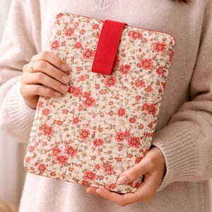 May include: Floral patterned tablet case with a red strap closure. The case features a cream background with a repeating pattern of red and pink flowers. The case is held by a person wearing a beige sweater.