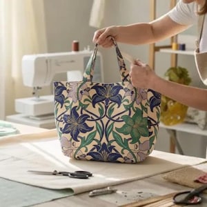 May include: A patterned tote bag featuring blue, green, and purple lily designs on a cream backdrop. The bag has two handles and is being held. A sewing machine and sewing supplies are visible in the background.