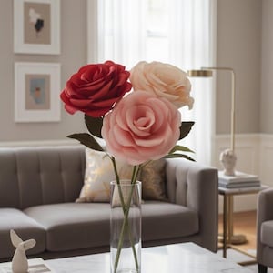 May include: A clear glass vase holding three large paper roses. The roses are in shades of red, peach, and pink, with green paper leaves. The vase sits on a white marble coffee table in a living room setting.