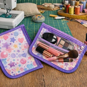 Puede incluir: Dos neceseres de cosméticos abiertos, con ribetes morados y frente transparente, uno lleno de maquillaje y brochas. El otro neceser está vacío, con un estampado pastel de corazones, estrellas y círculos. Los neceseres están sobre una superficie de madera.