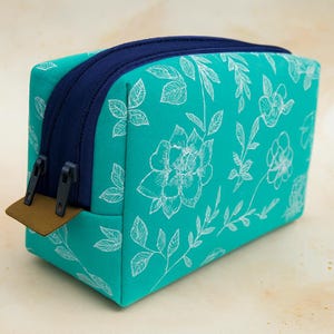 Könnte beinhalten: Türkisfarbene, rechteckige Kosmetiktasche mit weißem Blumenmuster. Die Tasche hat einen marineblauen Reißverschluss und Besatz sowie eine kleine braune Lasche an der Seite. Die Tasche ist für Make-up und andere Schönheitsprodukte konzipiert.
