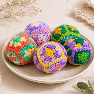 Puede incluir: Cinco bolas de ganchillo coloridas descansan sobre un plato blanco. Las bolas presentan patrones geom&eacute;tricos en tonos naranja, verde, morado, amarillo y rosa. Un peque&ntilde;o jarr&oacute;n beige y ramitas de flores secas est&aacute;n en el fondo.