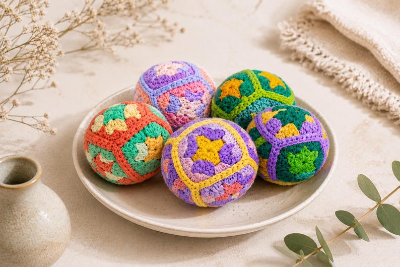 Puede incluir: Cinco bolas de ganchillo coloridas descansan sobre un plato blanco. Las bolas presentan patrones geom&eacute;tricos en tonos naranja, verde, morado, amarillo y rosa. Un peque&ntilde;o jarr&oacute;n beige y ramitas de flores secas est&aacute;n en el fondo.