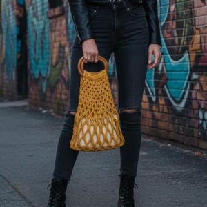 Peut inclure: Sac fourre-tout en crochet jaune moutarde avec des poign&eacute;es en bois. Le sac a un motif ajour&eacute; et une doublure cr&egrave;me. Le sac est tenu devant un mur de briques recouvert de graffitis.