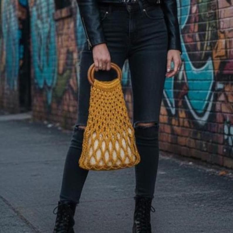 Peut inclure: Sac fourre-tout en crochet jaune moutarde avec des poign&eacute;es en bois. Le sac a un motif ajour&eacute; et une doublure cr&egrave;me. Le sac est tenu devant un mur de briques recouvert de graffitis.