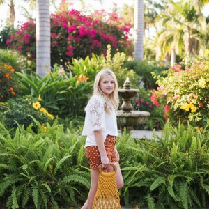 Peut inclure: Une personne porte un haut en dentelle blanc, un short marron &agrave; pois et des sandales, tenant un sac en macram&eacute; jaune avec des poign&eacute;es en bois. L'arri&egrave;re-plan pr&eacute;sente une v&eacute;g&eacute;tation luxuriante, des fleurs color&eacute;es et une fontaine.