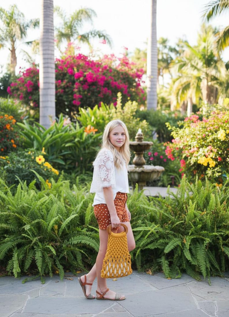 Peut inclure: Une personne porte un haut en dentelle blanc, un short marron &agrave; pois et des sandales, tenant un sac en macram&eacute; jaune avec des poign&eacute;es en bois. L'arri&egrave;re-plan pr&eacute;sente une v&eacute;g&eacute;tation luxuriante, des fleurs color&eacute;es et une fontaine.