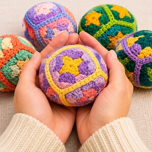 Puede incluir: Unas manos sostienen una colorida bola de ganchillo, rodeada de otras cuatro. Las bolas presentan patrones geom&eacute;tricos en tonos morado, amarillo, verde, naranja y azul. La persona lleva un su&eacute;ter color crema.