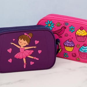 Op de afbeelding: Twee kleine, rechthoekige portemonnees. Eén is paars met een ballerina-ontwerp en roze hartjes. De andere is roze met een vogel, bloemen en cupcakes. Beide hebben ritsen en schouderbanden.