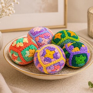 Puede incluir: Un cuenco decorativo contiene seis esferas de ganchillo coloridas. Cada esfera presenta un patr&oacute;n geom&eacute;trico &uacute;nico en tonos de naranja, verde, morado y amarillo. El cuenco tiene un borde dorado y est&aacute; sobre una superficie neutra.