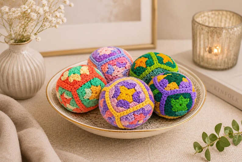 Puede incluir: Un cuenco decorativo contiene seis esferas de ganchillo coloridas. Cada esfera presenta un patr&oacute;n geom&eacute;trico &uacute;nico en tonos de naranja, verde, morado y amarillo. El cuenco tiene un borde dorado y est&aacute; sobre una superficie neutra.
