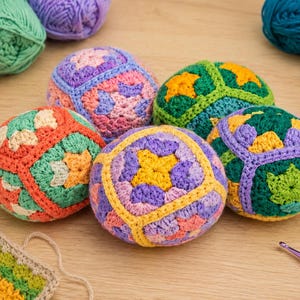 Puede incluir: Una colecci&oacute;n de bolas de ganchillo coloridas en varios tonos de naranja, verde, morado y amarillo. Las bolas est&aacute;n dispuestas sobre una superficie de madera clara, con madejas de hilo en colores a juego. Tambi&eacute;n se ven un ganchillo y una peque&ntilde;a muestra de ganchillo.