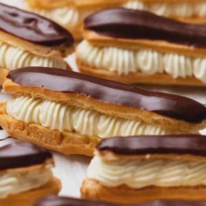 May include: Close-up of several eclairs. The pastries have a golden-brown choux pastry base, filled with white cream, and topped with a dark chocolate glaze. The arrangement highlights the textures and colours of the dessert.