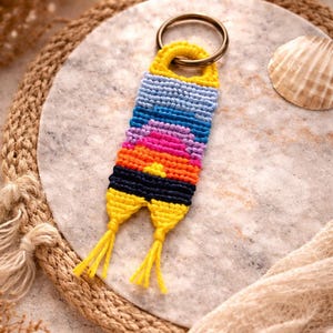 Op de afbeelding: Een handgemaakte macram&eacute; sleutelhanger met een zonsondergangontwerp. De sleutelhanger heeft een gele bovenkant met een metalen ring, overgaand in blauw, paars, oranje en zwart, met gele kwastjes aan de onderkant. Het ligt op een marmeren oppervlak.