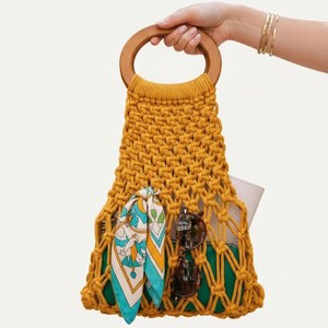 Peut inclure: Sac fourre-tout en macram&eacute; jaune moutarde avec une poign&eacute;e en bois. Le sac contient un foulard &agrave; motifs, des lunettes de soleil et une pochette blanche. Le sac est tenu par une personne avec des bracelets dor&eacute;s.