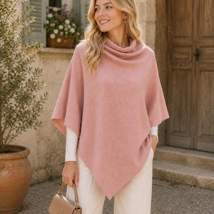 Könnte beinhalten: Ein staubrosa Poncho mit Rollkragen, getragen über einem weißen Langarmshirt. Der Poncho hat einen drapierten, asymmetrischen Saum. Die Person hält eine beige Handtasche und trägt eine weiße Hose. Der Hintergrund zeigt ein Steingebäude und eine Holztür.