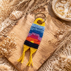 Op de afbeelding: Een handgemaakte macram&eacute; sleutelhanger met een kleurrijk zonsondergangontwerp. De sleutelhanger heeft een messing ring en gele kwastjes. Het kleurenschema omvat blauw, oranje, roze en geel.