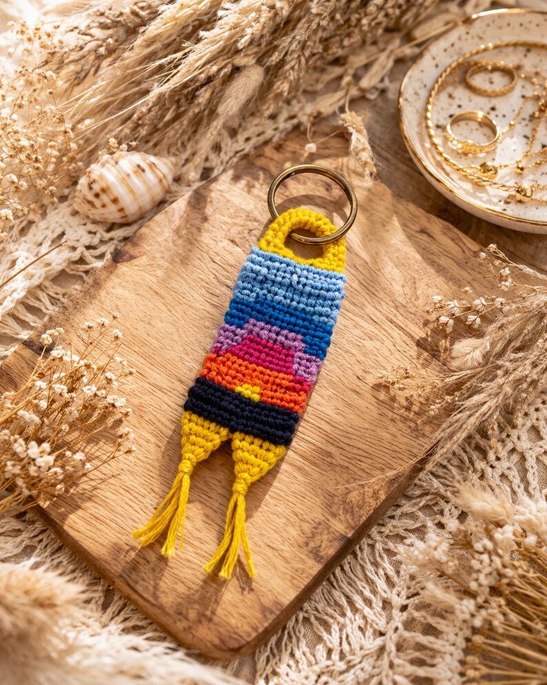 Op de afbeelding: Een handgemaakte macram&eacute; sleutelhanger met een kleurrijk zonsondergangontwerp. De sleutelhanger heeft een messing ring en gele kwastjes. Het kleurenschema omvat blauw, oranje, roze en geel.