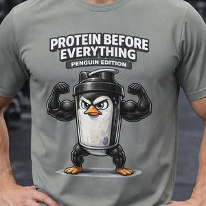 Op de afbeelding: Grijs t-shirt met de tekst "PROTEIN BEFORE EVERYTHING" en "PENGUIN EDITION". Het shirt toont een cartoonpinguïn met gespierde armen die een eiwitshake vasthoudt. De achtergrond is een sportschool.