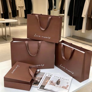 Puede incluir: Cuatro bolsas de papel marrón para la compra con asas de cinta marrón a juego. Cada bolsa presenta el texto cursivo blanco "Love myself" impreso en la parte frontal. Las bolsas están dispuestas en grupo sobre una superficie blanca junto a dos revistas.
