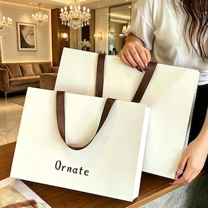 May include: Two rectangular white paper shopping bags with dark brown ribbon handles. The smaller bag in the front features the word Ornate printed in a simple black sans-serif font. Both bags have a smooth matte finish and a clean design.