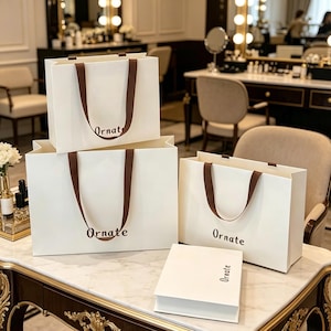 May include: Three off-white shopping bags and one rectangular off-white box. Each item features the word Ornate printed in brown. The bags have brown ribbon handles. The items are arranged on a white marble surface with gold-toned trim.