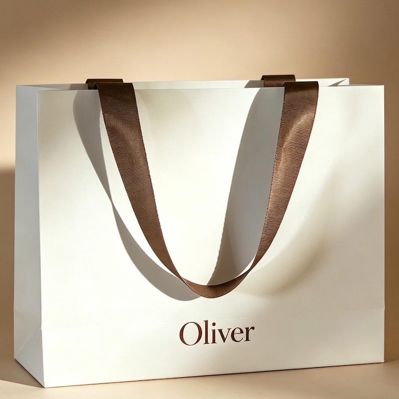 May include: A rectangular white paper shopping bag featuring the name Oliver printed in dark brown lettering on the front. The bag includes two dark brown ribbon handles with a textured finish, attached to the top edge of the bag.