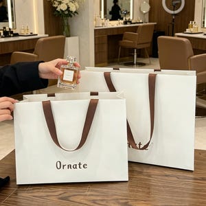 May include: A square glass perfume bottle with a gold cap and a white label, positioned next to two white paper shopping bags with brown ribbon handles. Each bag features the word Ornate printed in a dark brown font on the front surface.