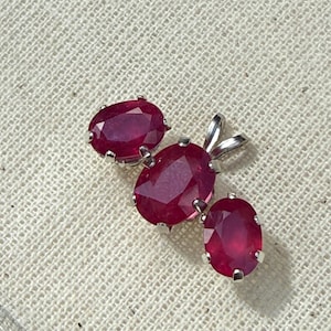May include: A pendant with three oval-cut, deep red gemstones set in silver. The central gem is larger, with two smaller gems flanking it. The gems have a faceted appearance, catching the light. The pendant is displayed on a textured, off-white surface.