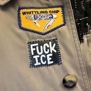May include: Close-up of a tan jacket adorned with patches. One patch reads "WHITTLING CHIP" with a knife illustration on a yellow background. Another patch says "FUCK ICE" in white letters on a black background. A brown button is visible.
