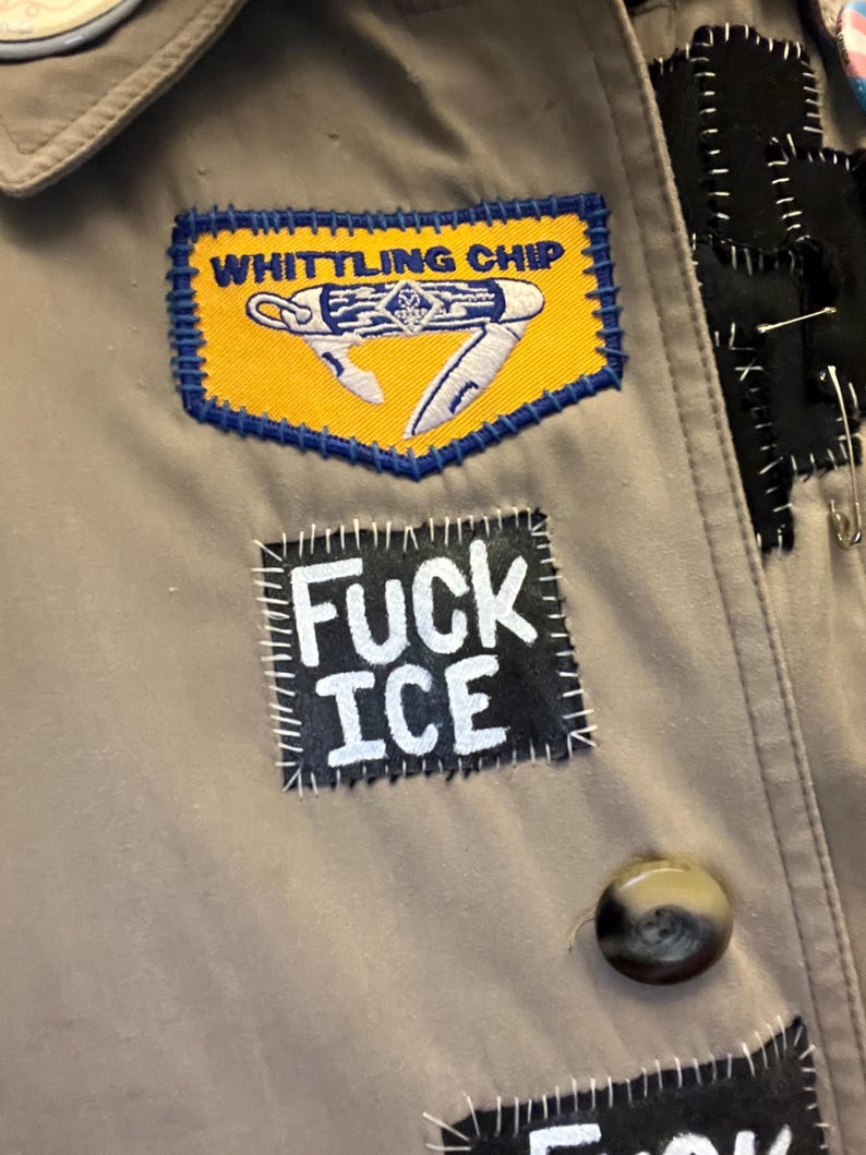 May include: Close-up of a tan jacket adorned with patches. One patch reads "WHITTLING CHIP" with a knife illustration on a yellow background. Another patch says "FUCK ICE" in white letters on a black background. A brown button is visible.