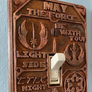 May include: A copper-colored light switch cover with embossed Star Wars-themed designs and text. The text includes "MAY THE FORCE BE WITH YOU!" and references to the Light Side, Dark Side, and Sith Lord. The switch is in the "ON" position.