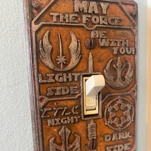 May include: A decorative light switch cover with a bronze finish. The cover features the text "MAY THE FORCE BE WITH YOU!" and "LIGHT SIDE", "DARK SIDE", "SITH LORD". The switch is in the "ON" position.