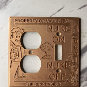 May include: A copper-colored rectangular light switch and outlet wall plate featuring embossed text that reads PROPERTY OF VAULT-TEC, NUKE ON, NUKE OFF, and FLIP RESPONSIBLY. The design includes a cartoon character, a mushroom cloud, and a rocket.