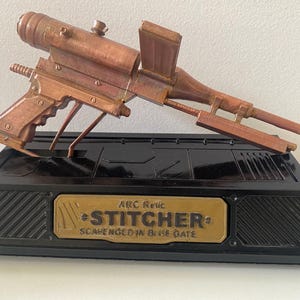 May include: A copper-colored, futuristic-looking prop gun, the "Stitcher," rests on a black base with a gold plaque. The gun features a pistol grip, a long barrel, and a top-mounted scope. The plaque reads "Stitcher" and "Scavenged in Blue Gate."