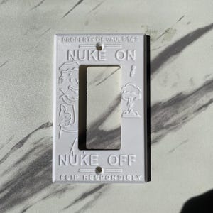 May include: A white rectangular light switch cover plate featuring embossed text and graphics. The top reads PROPERTY OF VAULT-TEC, NUKE ON, and the bottom reads NUKE OFF, FLIP RESPONSIBLY. A cartoon character is on the left and a mushroom cloud on the right.
