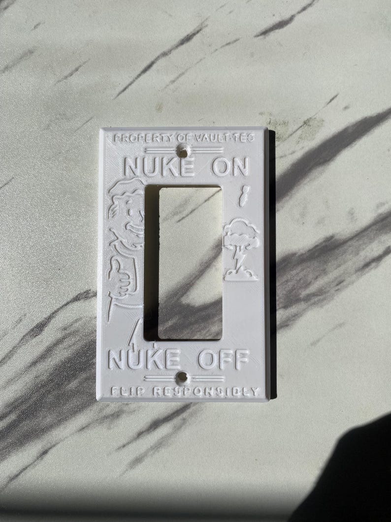 May include: A white rectangular light switch cover plate featuring embossed text and graphics. The top reads PROPERTY OF VAULT-TEC, NUKE ON, and the bottom reads NUKE OFF, FLIP RESPONSIBLY. A cartoon character is on the left and a mushroom cloud on the right.