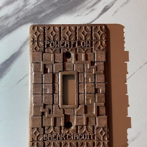 May include: A brown, textured light switch cover with the words "POWER FLOW", "BREAK CIRCUIT", and "MINECRAFT" embossed on the surface. The cover has a rectangular opening for the switch and two screw holes.