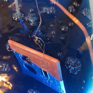 May include: A rectangular sign partially submerged in a deep blue liquid with visible bubbles. The sign features a copper-colored top section with the text PROPERTY OF VAULT-TEC and NUKE ON, and a blue bottom section with a rocket graphic.