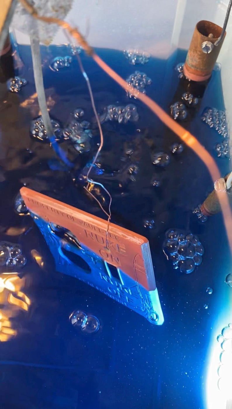 May include: A rectangular sign partially submerged in a deep blue liquid with visible bubbles. The sign features a copper-colored top section with the text PROPERTY OF VAULT-TEC and NUKE ON, and a blue bottom section with a rocket graphic.