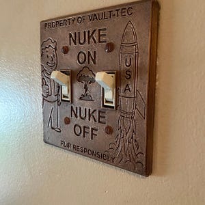 May include: A bronze-colored double light switch plate featuring the text PROPERTY OF VAULT-TEC, NUKE ON, NUKE OFF, and FLIP RESPONSIBLY. The plate includes an embossed cartoon character, a mushroom cloud, and a rocket labeled USA with two white toggles.