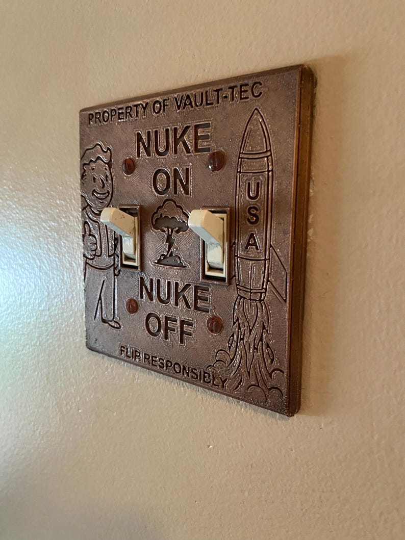 May include: A bronze-colored double light switch plate featuring the text PROPERTY OF VAULT-TEC, NUKE ON, NUKE OFF, and FLIP RESPONSIBLY. The plate includes an embossed cartoon character, a mushroom cloud, and a rocket labeled USA with two white toggles.